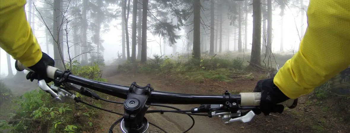 Photo taken from someone sitting on a mountain bike, of mountain bike handle bars and hands holding onto the handlebars, wearing a fluoro yellow top