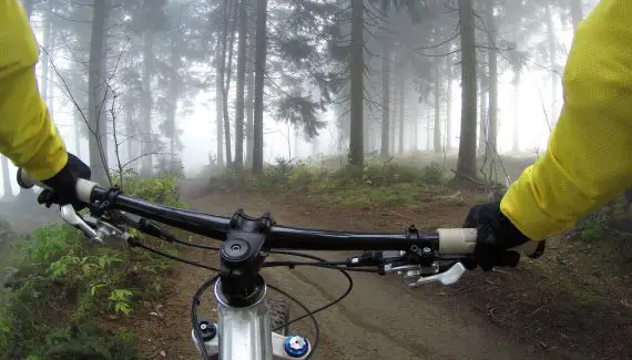 Photo taken from someone sitting on a mountain bike, of mountain bike handle bars and hands holding onto the handlebars, wearing a fluoro yellow top